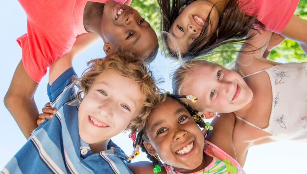 A diverse group of smiling children huddling together and looking down at the camera, symbolizing joy, connection, and the focus on student well-being.