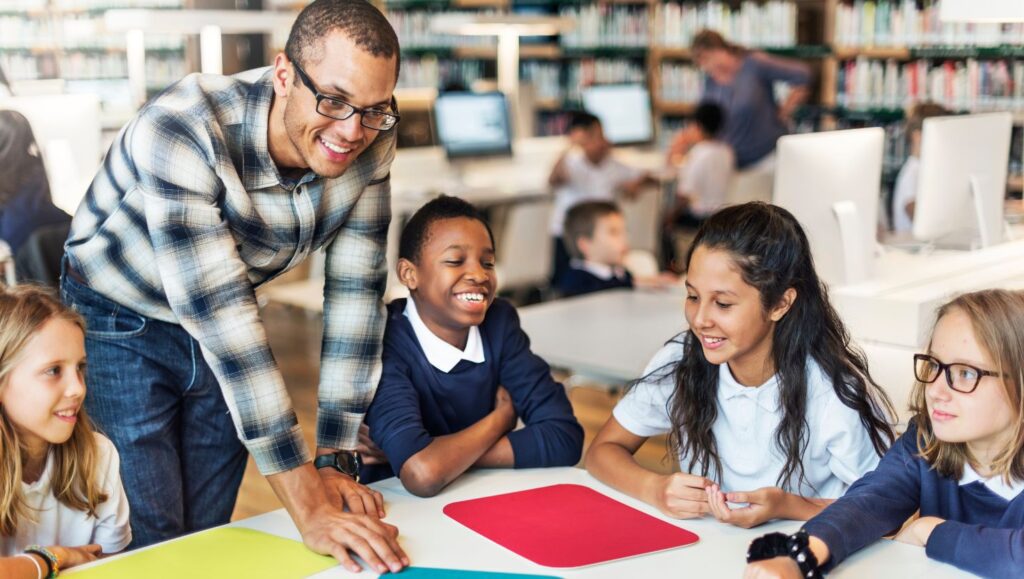 A smiling teacher interacting positively with diverse, engaged students at a table in a classroom, symbolizing strong teacher-student relationships and collaborative learning.